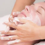 A woman with eyes closed receives a facial massage, with hands applying creamy lotion to her face. The scene conveys relaxation and self-care.