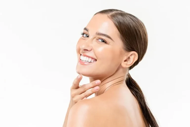 A woman with smooth skin and a bright smile looks back over her shoulder against a white background. Her hair is slicked back, conveying a fresh, joyful mood.