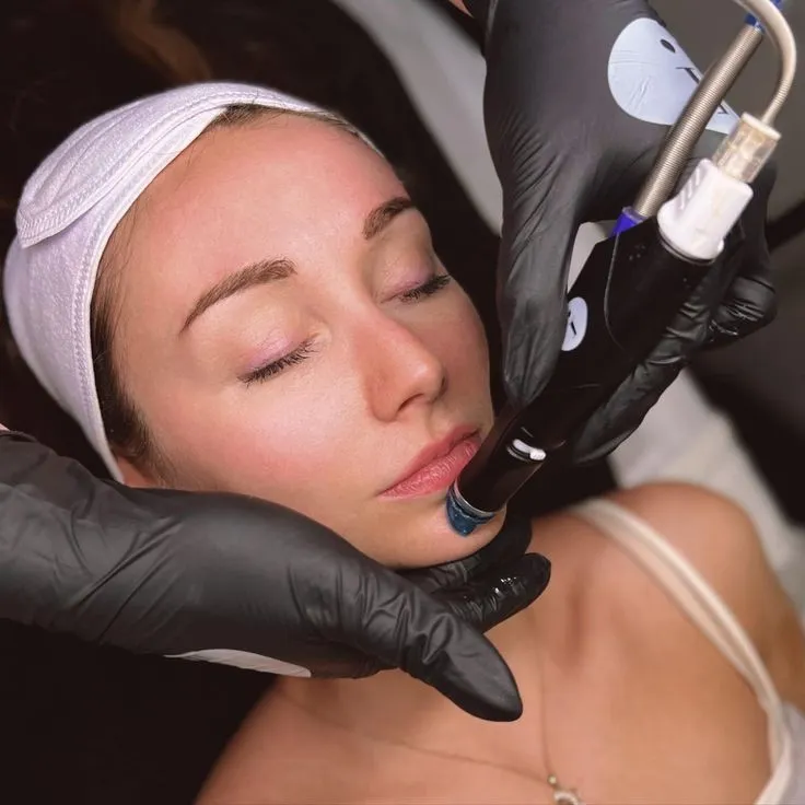 Woman receiving a facial treatment. A person in black gloves uses a skincare device on her chin. Her eyes are closed, and she appears relaxed.