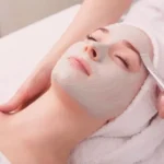 A person relaxes with a facial mask applied, eyes closed and head wrapped in a towel, while receiving a gentle face massage in a spa-like setting.