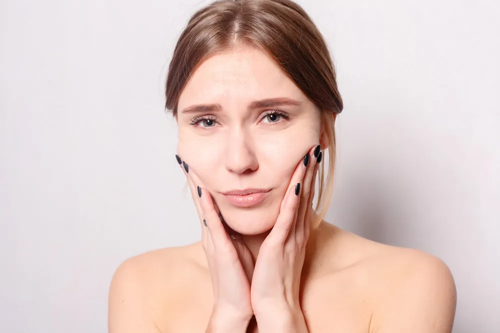 A worried woman with light brown hair touches her cheeks, her expression showing concern. She has dark nail polish and a neutral background.