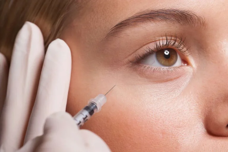 Close-up of a person’s face receiving a cosmetic injection near the eye. A gloved hand holds the syringe, conveying a clinical and careful tone.