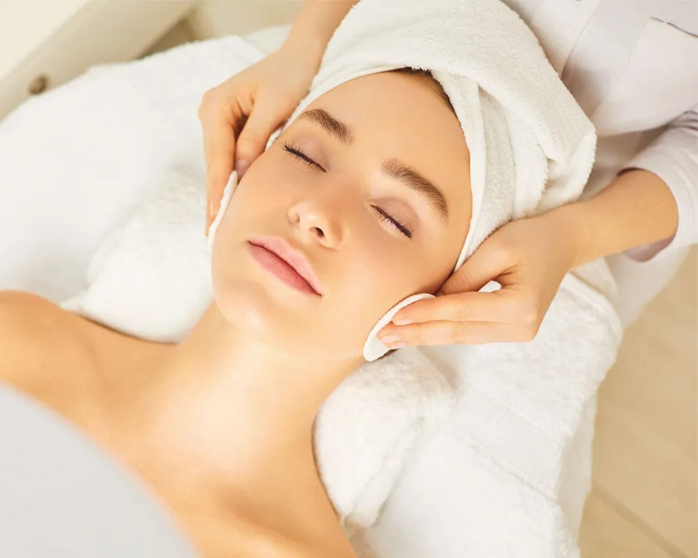 A person receives a relaxing facial treatment, eyes closed, with a towel wrapped around their head. Hands gently cleanse their face, conveying calmness.