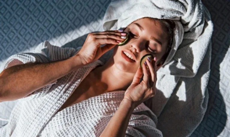 A woman in a white robe and towel turban relaxes on a patterned surface, eyes closed, smiling gently while applying cucumber slices to her eyes.
