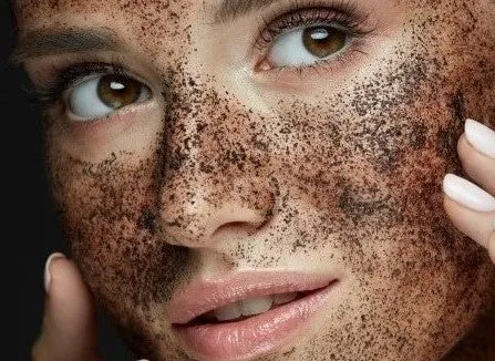A woman's face with a coffee scrub applied, displaying smooth skin and relaxed expression. Her hand gently touches her cheek, conveying a sense of calm.