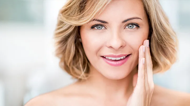 A woman with short blonde hair smiles gently, touching her cheek with her hand. She exudes happiness and confidence, set against a soft, blurred background.