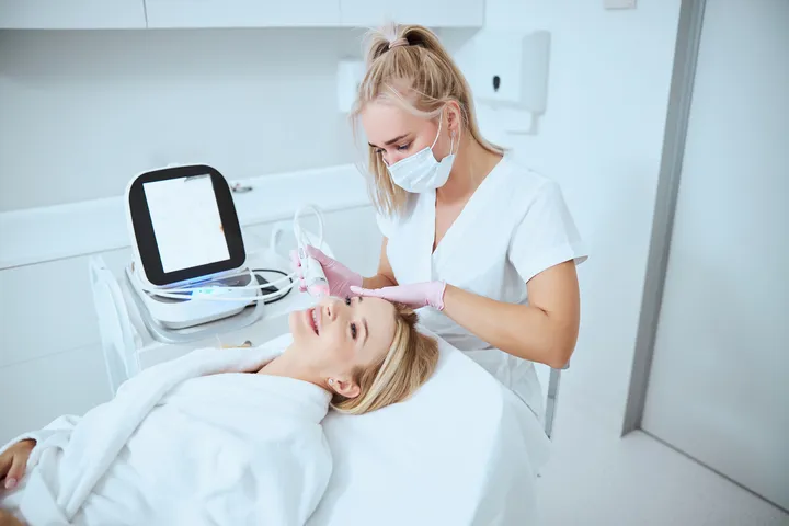 A woman in a spa, wearing a mask and gloves, performs a facial treatment on a smiling client in a white robe, using a skincare machine.