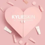 Pink heart with "Kylie Skin by Kylie Jenner" text in center, surrounded by various light pink skincare products on a pink background. Elegant and soothing tone.
