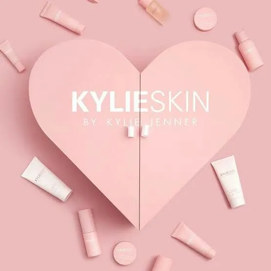 Pink heart with "Kylie Skin by Kylie Jenner" text in center, surrounded by various light pink skincare products on a pink background. Elegant and soothing tone.