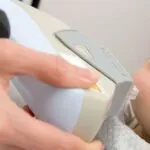 A hand holds a laser hair removal device against a person's skin, suggesting a cosmetic procedure. The setting appears clinical and professional.