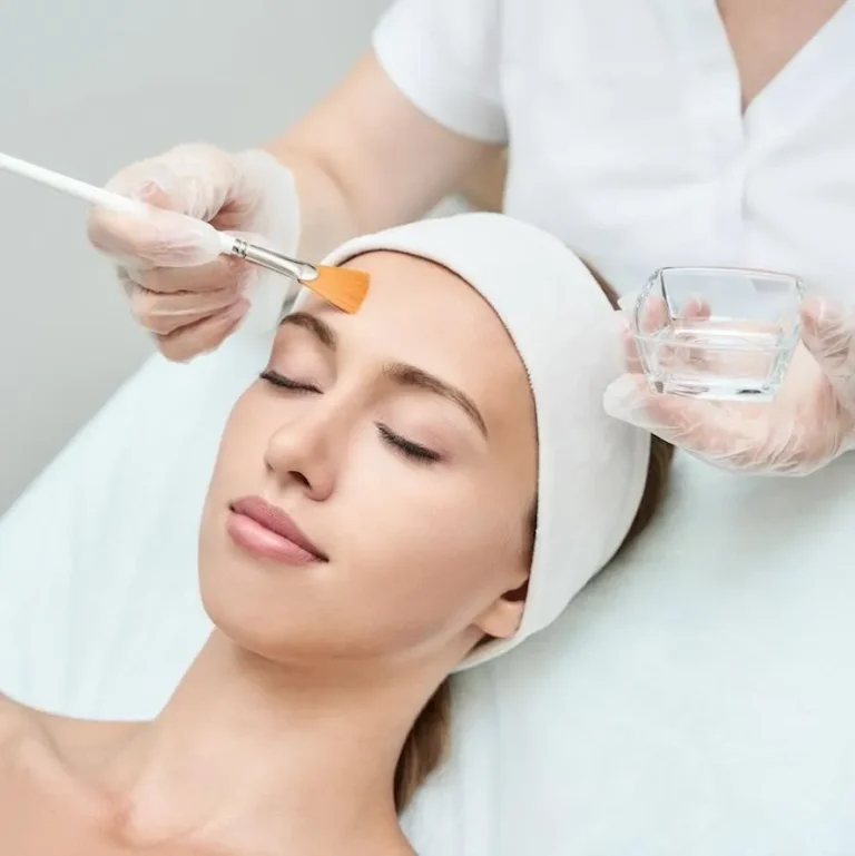 A woman with closed eyes, wearing a headband, receives a facial treatment with a brush. The setting is serene and professional, suggesting relaxation.