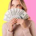 A person in a beige sweater holds a fanned-out stack of cash, partially covering their face. The background is split pink and yellow, conveying a playful tone.