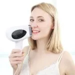 A smiling woman with blonde hair uses a handheld laser hair removal device on her face. She wears a light-colored top against a bright background.