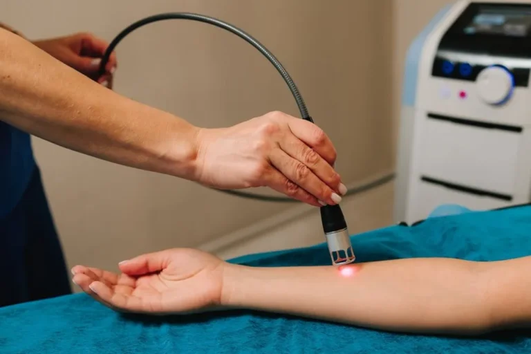 A person's arm receives laser therapy from a handheld device emitting red light, operated by another person's hands. The scene conveys a clinical, healing environment.