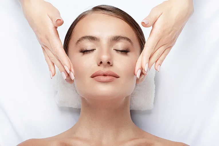 Woman receiving a relaxing facial massage with closed eyes, lying on a white towel. Hands gently touch her cheeks, conveying calmness and serenity.